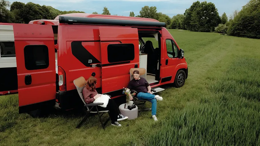 Bürstner Papillon Der Büstner Papillon statisch von schräg hinten rechts auf einer Wiese. Zwei Personen sitzen auf Campingstühlen vor der geöffneten Schiebetüre.