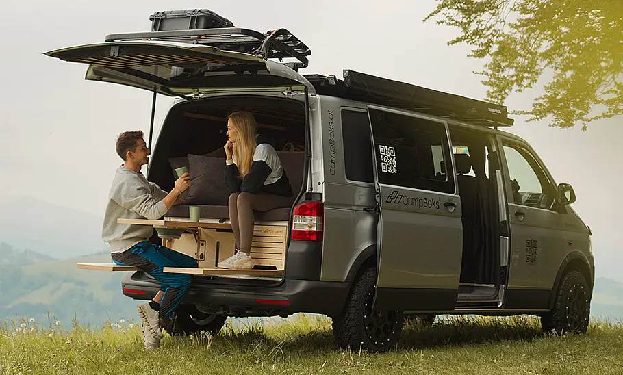 Das Bild zeigt das CampBoks Campingbox-Modul im Heck eines VW T6. Eine Person sitzt auf dem Bett, eine weitere auf der ausfahrbaren Sitzbank.