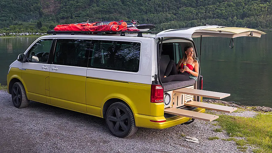 Eine CampBoks Campingbox in einem VW T6.1 mit geöffneter Heckklappe, der von schräg hinten links an einem See parkt. Eine Person sitzt im Heckbereich auf dem Bett.