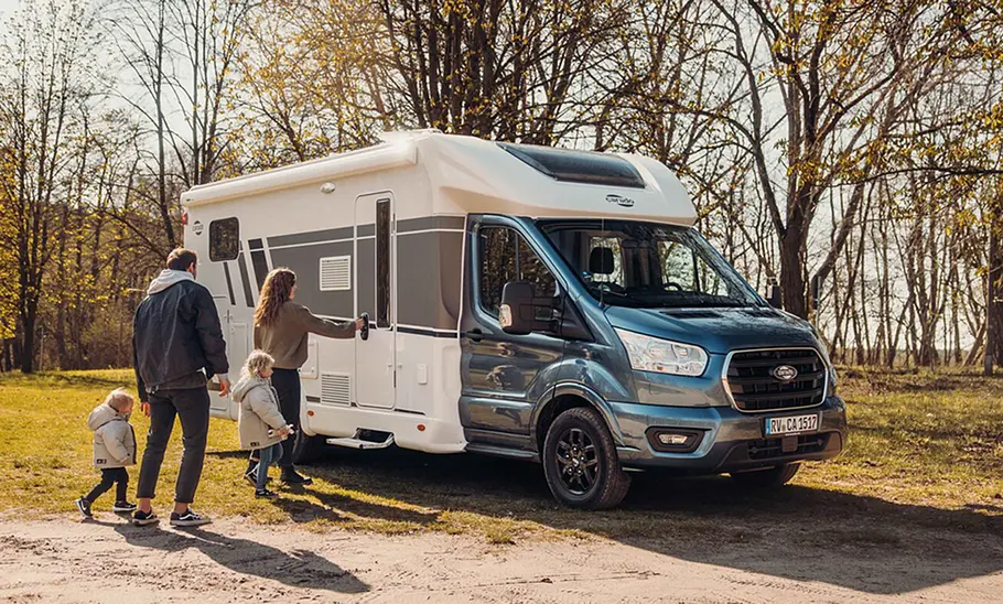Carado T447 (2024) Das Bild zeigt den Carado T447 Edition25 (2024) auf Basis des Ford Transit stehend von schräg vorne rechts fotografiert. Eine Familie läuft auf die Aufbautüre des Wohnmobils zu.