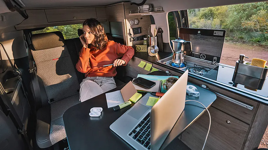 Ansicht des Wohnraums im Citroën Holidays. Im Fokus ist die Rückbank mit Person am Tisch, Laptop auf dem Tisch, Espressokocher auf dem Gasherd.