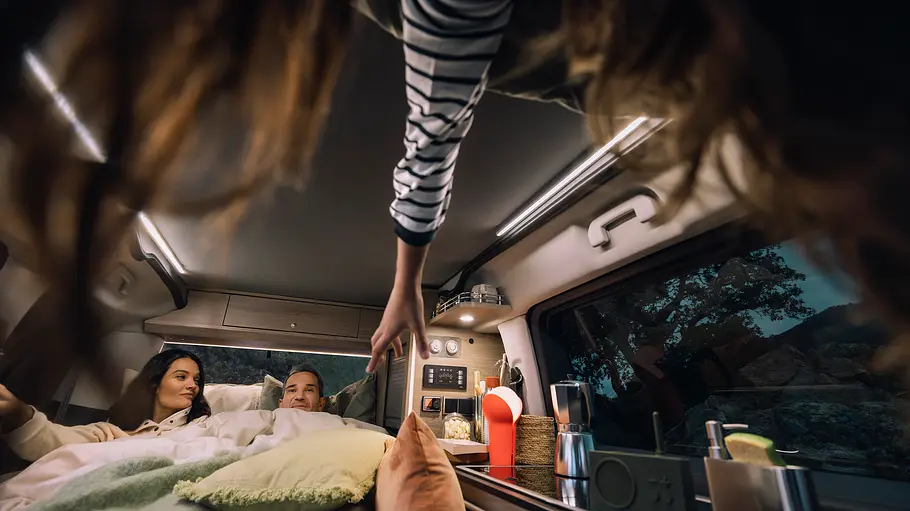 Der Wohnraum des Citroën Holidays bei Nacht. Im unteren Bett liegen zwei Erwachsene, ein Kind schaut durch die Dachöffnung vom Schlafdach herunter und streckt die Hand nach einem Kissen aus.