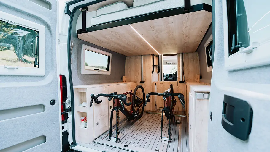 Clever Vans Runner 636 Active Die Aufnahme von außen nach innen zeigt das bis unters Dach hochgezogene Hubbett und die darunter liegenden Heckgarage bei geöffneten Seitentüren.
