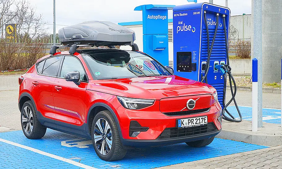 Das Bild zeigt einen Volvo C40 Recharge Pure Electric an einer Ladesäule für Elektroautos von schräg vorne rechts fotografiert. Eine Dachbox ist auf Dachträgern angebracht.