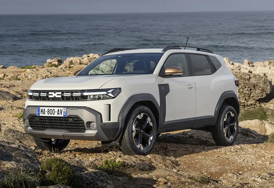 Dacia Duster Leasing Der Dacia Duster in Creme-Weiß statisch von schräg vorn links vor einem felsigen Küstenabschnitt abgestellt.