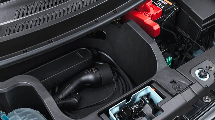 Ein Ladekabel liegt aufgerollt in einem Staufach unter der Fronthaube des Dacia Spring, neben einer 12-V-Batterie mit roter Polabdeckung.