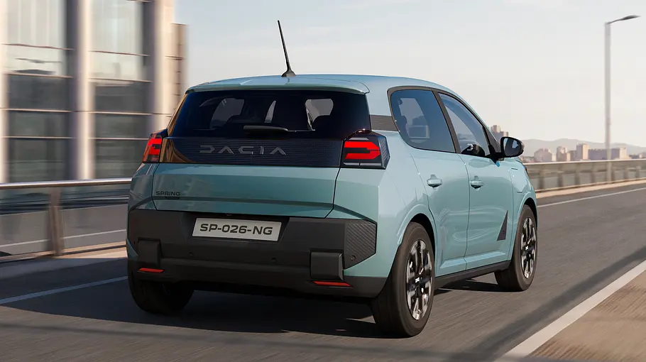 Ein hellblauer Dacia Spring fährt auf einer mehrspurigen Straße. Das Auto ist schräg von hinten vor einer Stadtkulisse zu sehen.