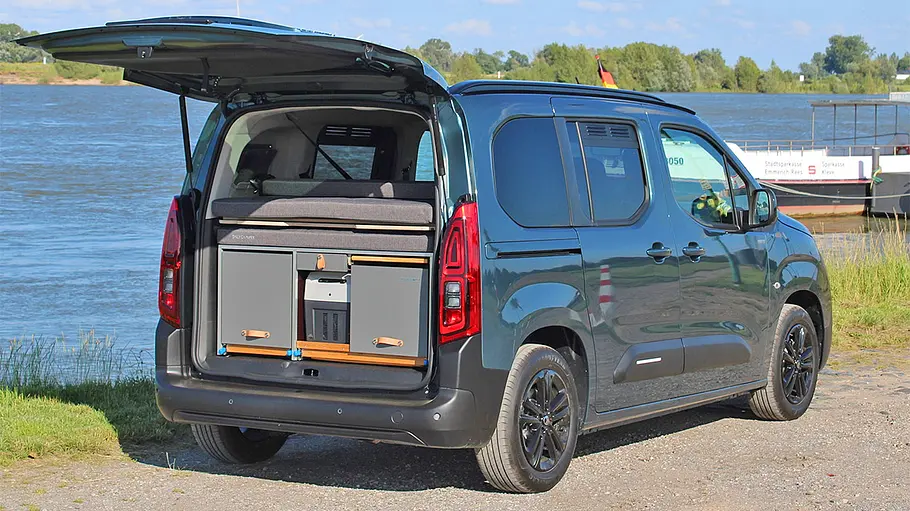 Die Campingbox Dailycamper Trekking in einem stehenden Fiat Doblo mit geöffneter Heckklappe. Das Fahrzeug ist von schräg hinten rechts gezeigt.