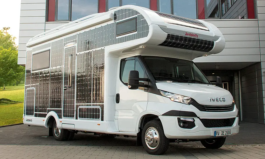 Dethleffs E.Home (2017) steht vor einem Geschäftsgebäude, die Seiten des Wohnmobils sind flächendeckend mit Solarfolie beklebt, Ansicht von vorne rechts.