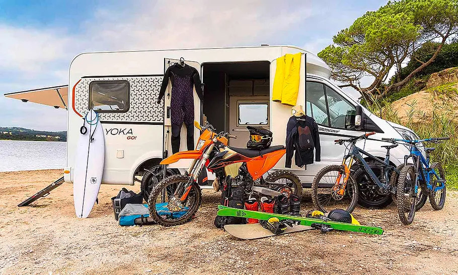 Dethleffs Yoka Go T1 (2024) Das Bild zeigt den Dethleffs Yoka Go T1 (2024) an einem Strand stehend mit geöffneter Heckklappe und geöffneter Seitentüre von rechts fotografiert. Vor dem Camper steht Sportequipment, wie Mountainbikes, ein Motocross-Motorrad und Surfbretter.