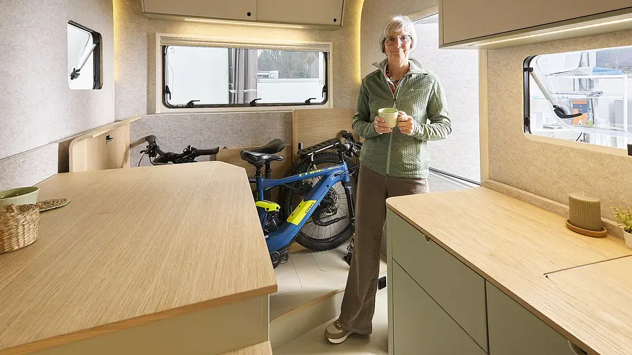Die Innenaufnahme zeigt Fahrräder die in der die zusammengeklappte Sitzgruppe der Dethleffs-Studie c.fold Platz finden. Davor steht eine Frau mit Kaffeetasse.