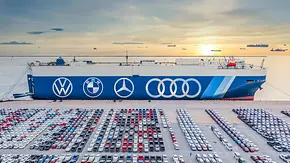 Symbolbild zeigt Autotransporter angedockt am Hafen, davor Neuwagen, die verfrachtet werden sollen. Die Flanke des Schiffes ziert die Markenlogos von VW, BMW, Mercedes und Audi. - Foto: Adobe Stock/AUTO ZEITUNG