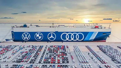 Symbolbild zeigt Autotransporter angedockt am Hafen, davor Neuwagen, die verfrachtet werden sollen. Die Flanke des Schiffes ziert die Markenlogos von VW, BMW, Mercedes und Audi. - Foto: Adobe Stock/AUTO ZEITUNG