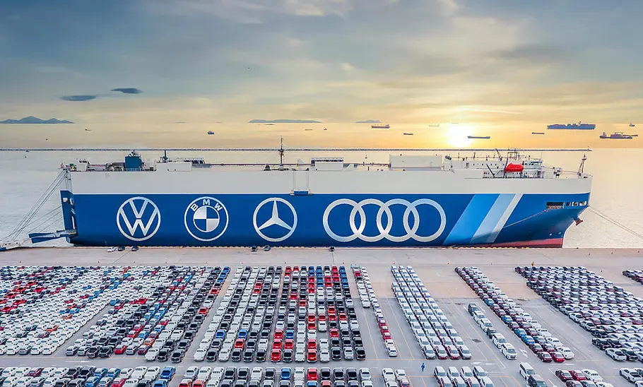 Symbolbild zeigt Autotransporter angedockt am Hafen, davor Neuwagen, die verfrachtet werden sollen. Die Flanke des Schiffes ziert die Markenlogos von VW, BMW, Mercedes und Audi.