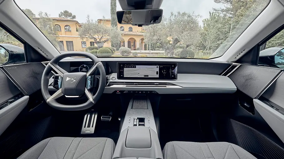 DS No. 7 im Fahrbericht Der Innenraum des DS No. 7 mit Lenkrad, breitem Display und grauen Sitzen. Durch die Frontscheibe sind ein Haus und Bäume zu sehen.