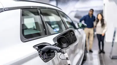 Ein E-Auto, das gerade geladen wird und dahinter sieht man leicht verschwommen ein Paar, das auf das Fahrzeug blickt. - Foto: Imago