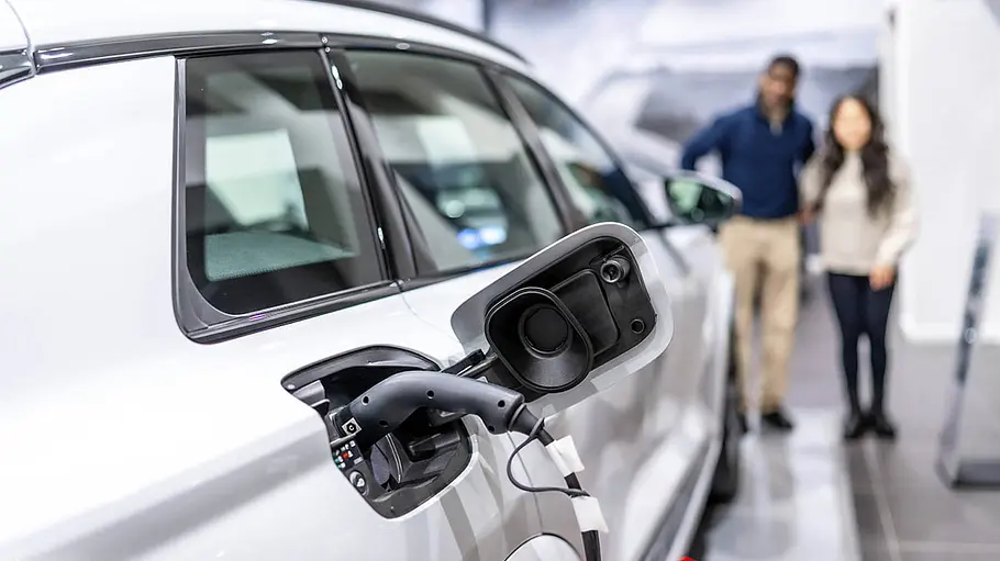 E-Auto kaufen oder leasen? Ein E-Auto, das gerade geladen wird und dahinter sieht man leicht verschwommen ein Paar, das auf das Fahrzeug blickt.