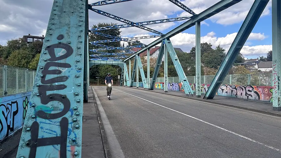 E-Scooter Segway Ninebot F3 Pro D Die Außenaufnahme zeigt einen Testredakteur auf dem E-Scooter Segway Ninebot F3 Pro D von Weitem mit eingeschaltetem Licht auf einer Eisenbahnbrücke.