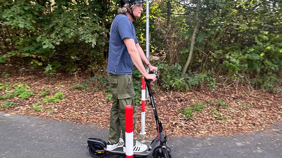 E-Scooter Segway Ninebot F3 Pro D Die Außenaufnahme zeigt einen Testredakteur auf dem E-Scooter Segway Ninebot F3 Pro D, der auf einem Fahrradweg fährt, beim Durchqueren von zwei Begrenzungspfählen.
