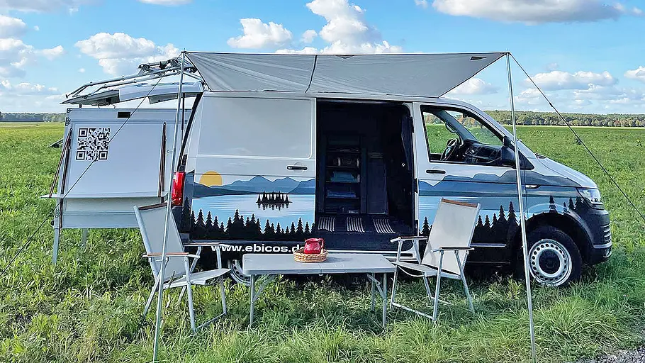 Das Ebicos Happy Dreamer in einem VW T6.1. Das Fahrzeug ist im Profil von rechts zu sehen, vor der geöffneten Schiebetüre stehen Tisch und Stühle, eine Markise ist über den Möbeln aufgespannt.