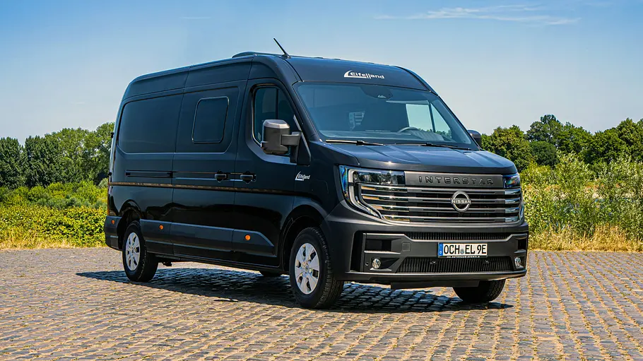 Der Eifelland Nissan Interstar-e steht auf einem gepflasterten Platz vor Sträuchern, fotografiert von schräg vorne rechts.