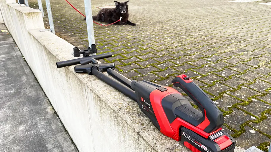 Der Akku-Handstaubsauger Einhell TE-HV 18/06 Li - Solo liegt mit Zubehör auf einer Betonstufe. Im Hintergrund ist ein Hund zu sehen.
