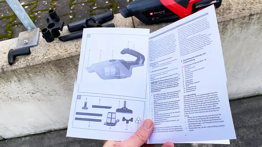 Eine Bedienungsanleitung in der Hand eines Testers. Dahinter liegt der Akku-Handstaubsauger Einhell TE-HV 18/06 Li – Solo auf einer Mauer.