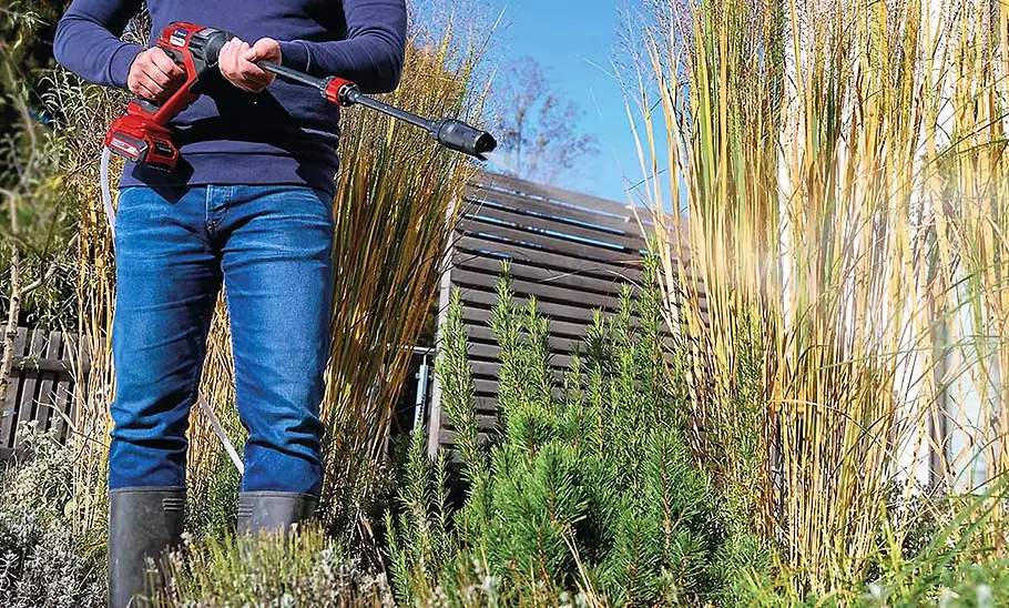 Mitteldruckreiniger Einhell Hypresso 18/24-1 im Test Eine Person steht in einem Garten, hat die Sprühpistole des Einhell Hypresso 18/24-1 in beiden Händen und sprüht Wasser.