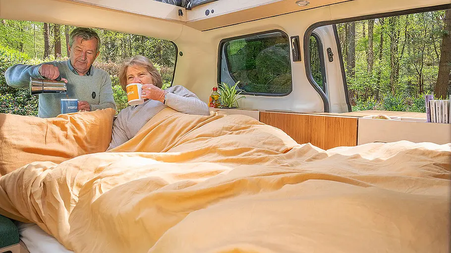 eVentje (2024); Innenraum, Ansicht auf den Wohnraum in Nachtstellung. Eine Person liegt im Bett, eine zweite steht hinter dem Van.