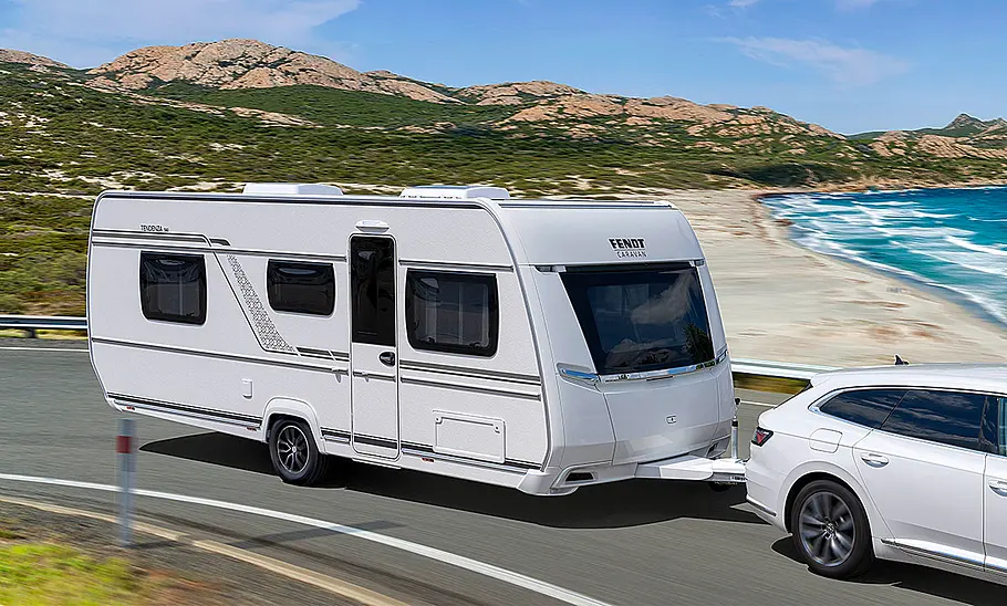 Das Bild zeigt den Fendt Tendenza (2025) von schräg vorne rechts fotografiert. Der Wohnwagen wird von einem VW Arteon an einem Stand vorbeigezogen.
