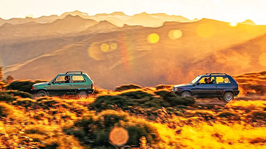 Blick von der Seite auf zwei Fiat Panda 4x4, die im Sonnenuntergang durch eine hügelige Landschaft fahren.