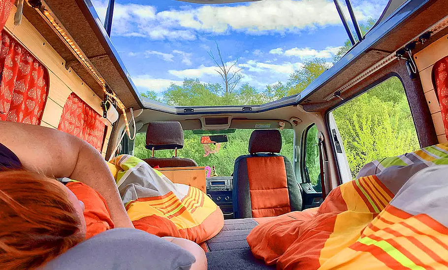 Flowcamper Frieda Das Bild zeigt den Wohnraum des Flowcamper Frieda im Bettumbau. Das Bild wurde vom Bettende aufgenommen, der Blick geht leicht nach oben durch das geöffnete Aufstelldach in den Himmel. Eine Person liegt auf dem Bett.