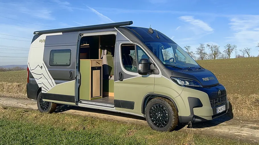 Flowcamper Moritz Der Flowcamper Moritz statisch von schräg vorne links mit geöffneter Schiebetüre auf einem Feldweg.