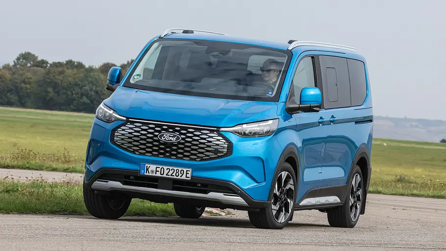 Der Ford E-Tourneo Custom fährt durch eine Rechtskurve, von vorne links fotografiert.