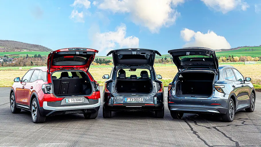 Ford Explorer/Kia EV5/MG S5 EV Blick von schräg hinten auf die drei kompakten E-SUV Ford Explorer, Kia EV5 und MG S5 EV mit geöffneten Kofferraumklappen.