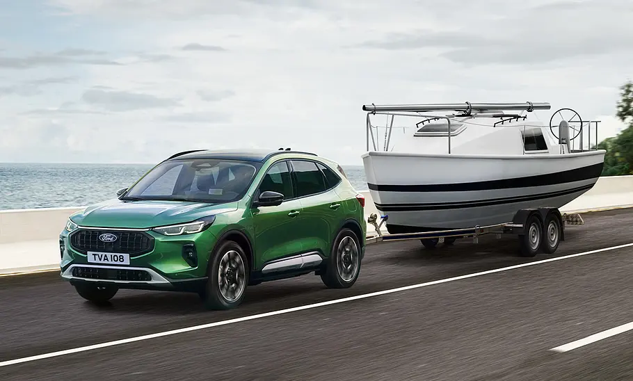 Das Ford Kuga Facelift (2024) zieht einen Anhänger mit einem Boot, von vorne links fotografiert.