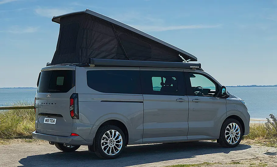 Die Außenaufnahme zeigt den Ford Transit Custom Nugget L2 (2025), der an der Küste parkt, in der Ansicht von schräg hinten rechts.