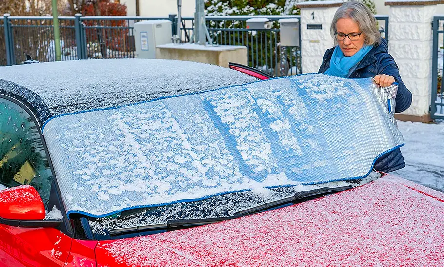 Eine Person hebt die Frontscheibenabdeckung von der Windschutzscheibe eines roten Audi A1 nach einer frostigen Nacht. Auf Auto und Abdeckung liegt etwas Schnee.