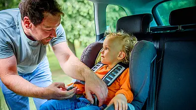 Das Bild zeigt eine Person, die durch die geöffnete Hintertüre auf der rechten Fahrzeugseite ein Kind in einem Kindersitz anschnallt. Ansicht vom Fahrersitz aus. - Foto: Imago