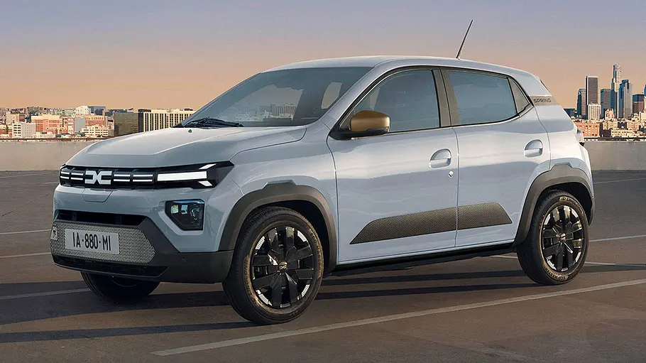Das Dacia Spring Facelift (2024) steht vor urbanem Hintergrund auf einem Parkplatz, von vorne links fotografiert.