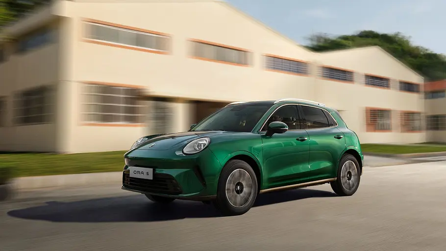 GWM-Ora-5-Hybrid-Fahrbericht Ein Ora 5 Hybrid in Grün fährt von schräg vorn betrachtet vor einem Haus entlang.