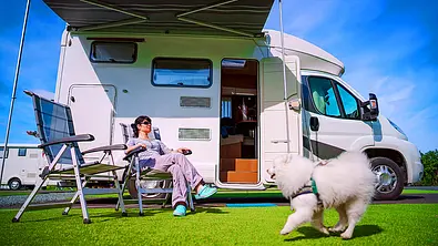 Das Bild zeigt einen kleinen weißen Hund, der an einem Wohnmobil auf einem Campingplatz vorbeirennt. Vor dem Wohnmobil sitzt unter der Markise eine Frau auf einem Campingstuhl. - Foto: Imago
