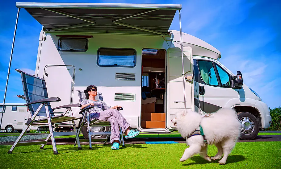 Das Bild zeigt einen kleinen weißen Hund, der an einem Wohnmobil auf einem Campingplatz vorbeirennt. Vor dem Wohnmobil sitzt unter der Markise eine Frau auf einem Campingstuhl.
