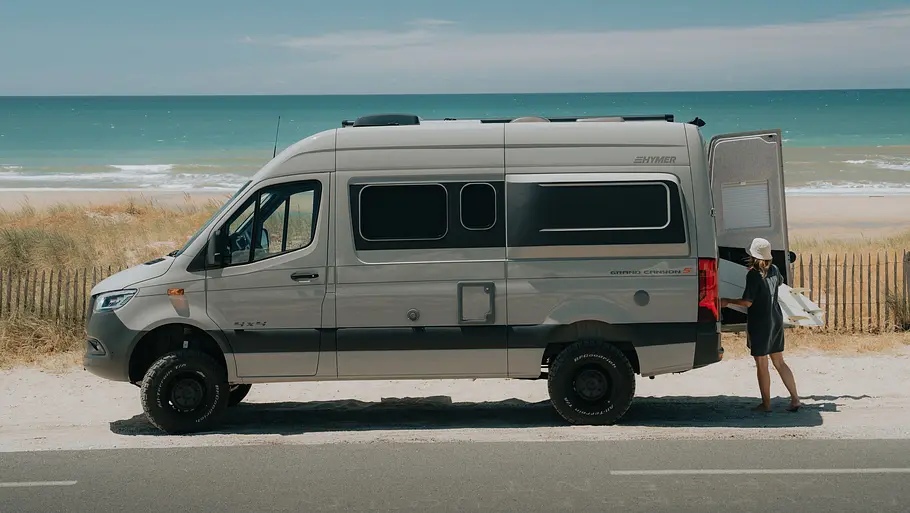 Der Hymer Grand Canyon S statisch von links am Straßenrand geparkt. Im Hintergrund ist ein Strand zu sehen, eine Person lädt Gepäck durch die geöffnete Hecktüre ins Fahrzeug.