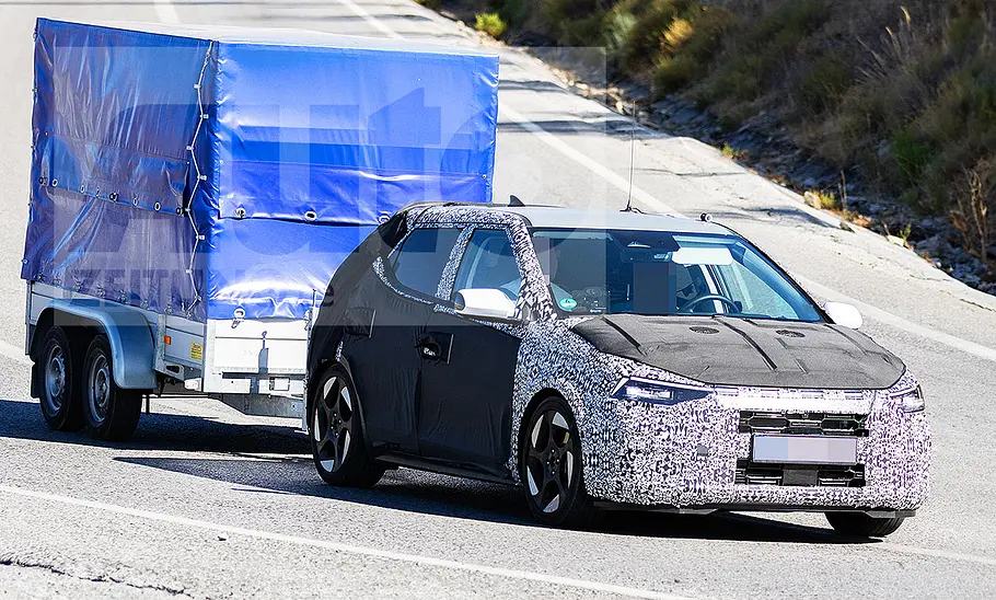 Blick von schräg vorne auf den fahrenden Erlkönig des Hyundai i20 (2026), der einen Anhänger zieht.