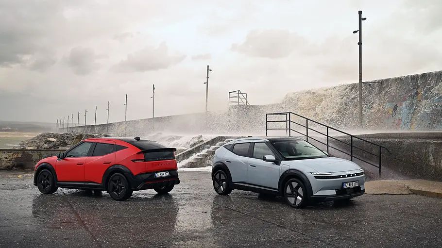 Zwei Hyundai Ioniq 3 stehen auf einer Asphaltfläche am Meer mit Gischt im Hintergrund. Ein Fahrzeug ist blau, das andere rot.