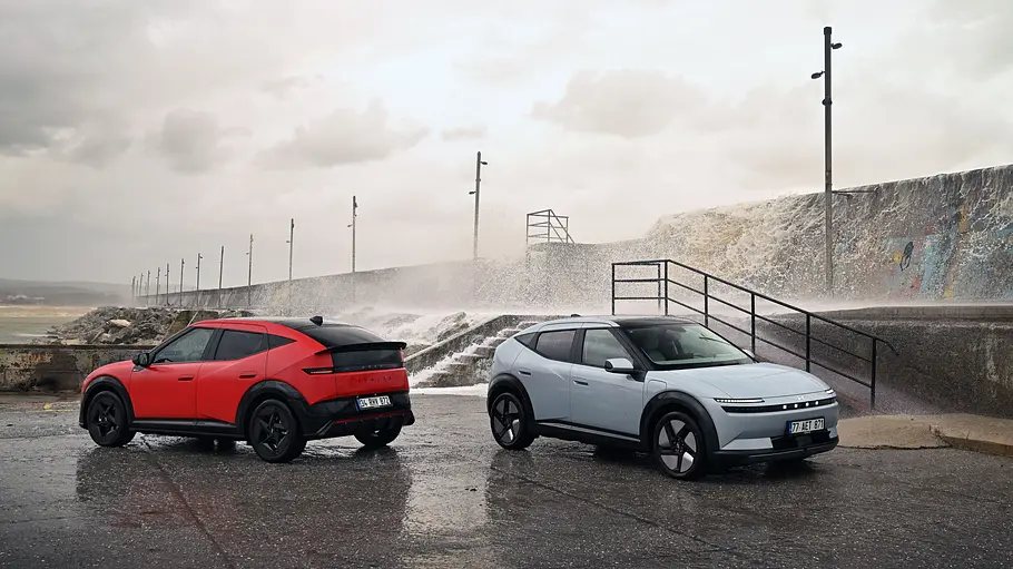 Zwei Hyundai Ioniq 3 stehen auf einer Asphaltfläche am Meer mit Gischt im Hintergrund. Ein Fahrzeug ist blau, das andere rot.