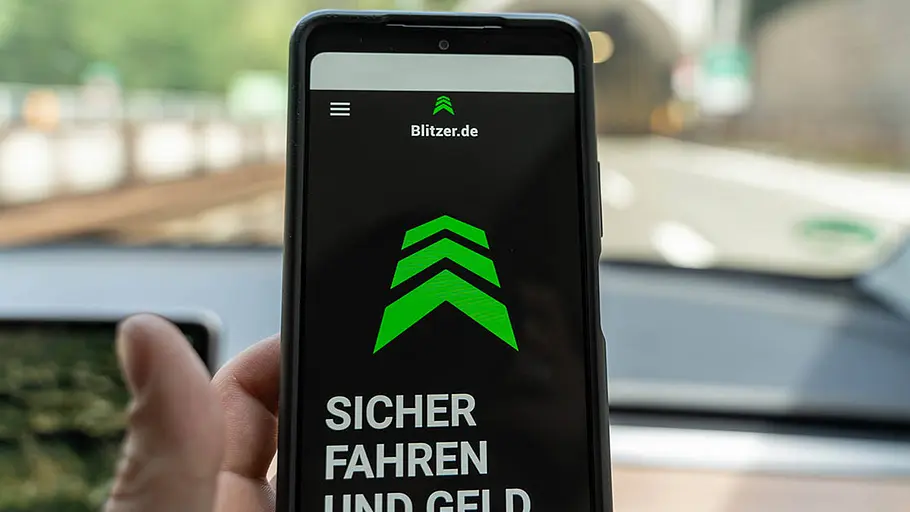 Ein Handydisplay zeigt einen Blitzerwarner an. In Hintergrund unscharf der Blick durch eine Auto-Frontscheibe auf die Straße.