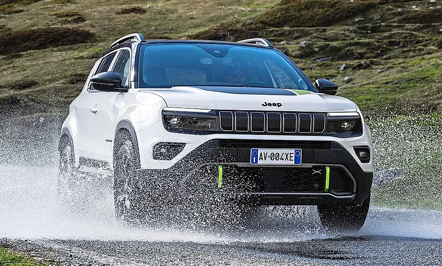 Jeep Avenger 4xe (2025) Der Jeep Avenger 4xe (2025) fährt durch eine Pütze und spritzt Wasser hoch. Von schräg vorne fotografiert.