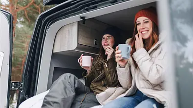Zwei junge Frauen im Outdoor-Outfit sitzen im Heck eines Campingbusses und trinken Kaffee. - Foto: Imago